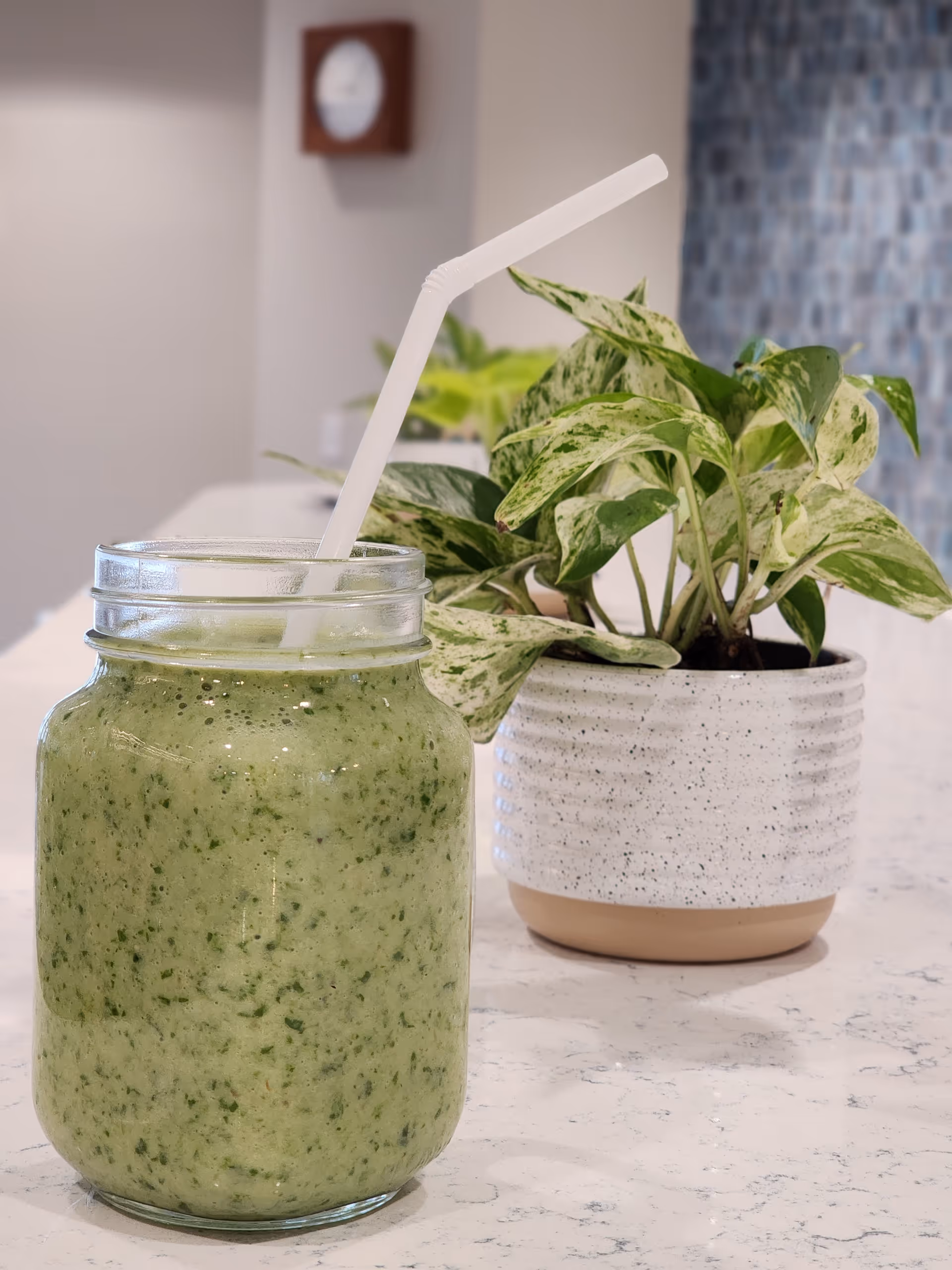 A glass jar filled with a green smoothie and a white bendable straw on a marble countertop, next to a white speckled ceramic pot containing a green and white variegated plant. The background shows a blurred wall clock and a tiled wall.
