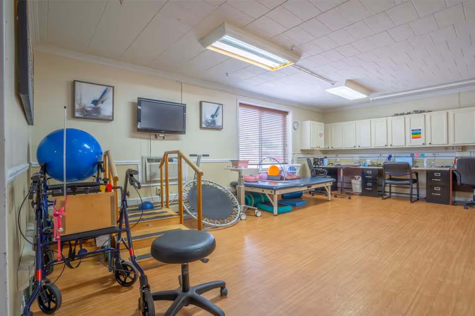 A spacious rehabilitation room with wooden flooring, featuring exercise equipment such as a walker, a large blue exercise ball, a small trampoline, parallel bars, and a therapy table with various therapy tools. The room has a wall-mounted TV, two framed pictures, a window with blinds, and a long counter with cabinets and office chairs underneath.
