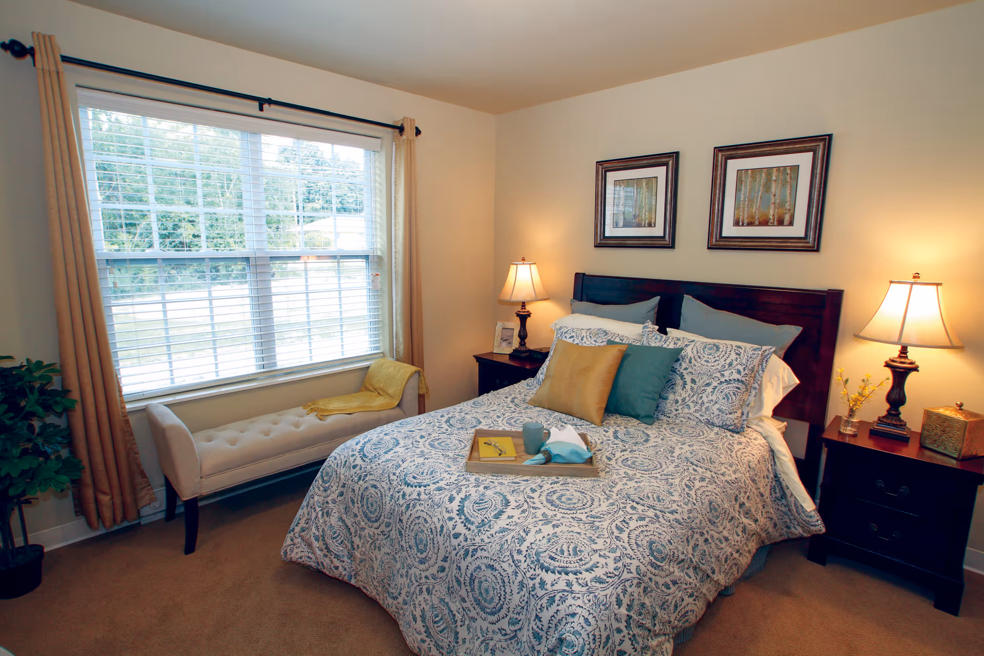 A cozy bedroom with a large window covered by beige curtains and white blinds. The bed has a patterned blue and white comforter, with three decorative pillows in yellow, teal, and blue. There are two dark wooden nightstands on either side of the bed, each with a lamp and decorative items. A beige cushioned bench with a yellow throw blanket is placed under the window, and a small green plant is in the corner of the room.
