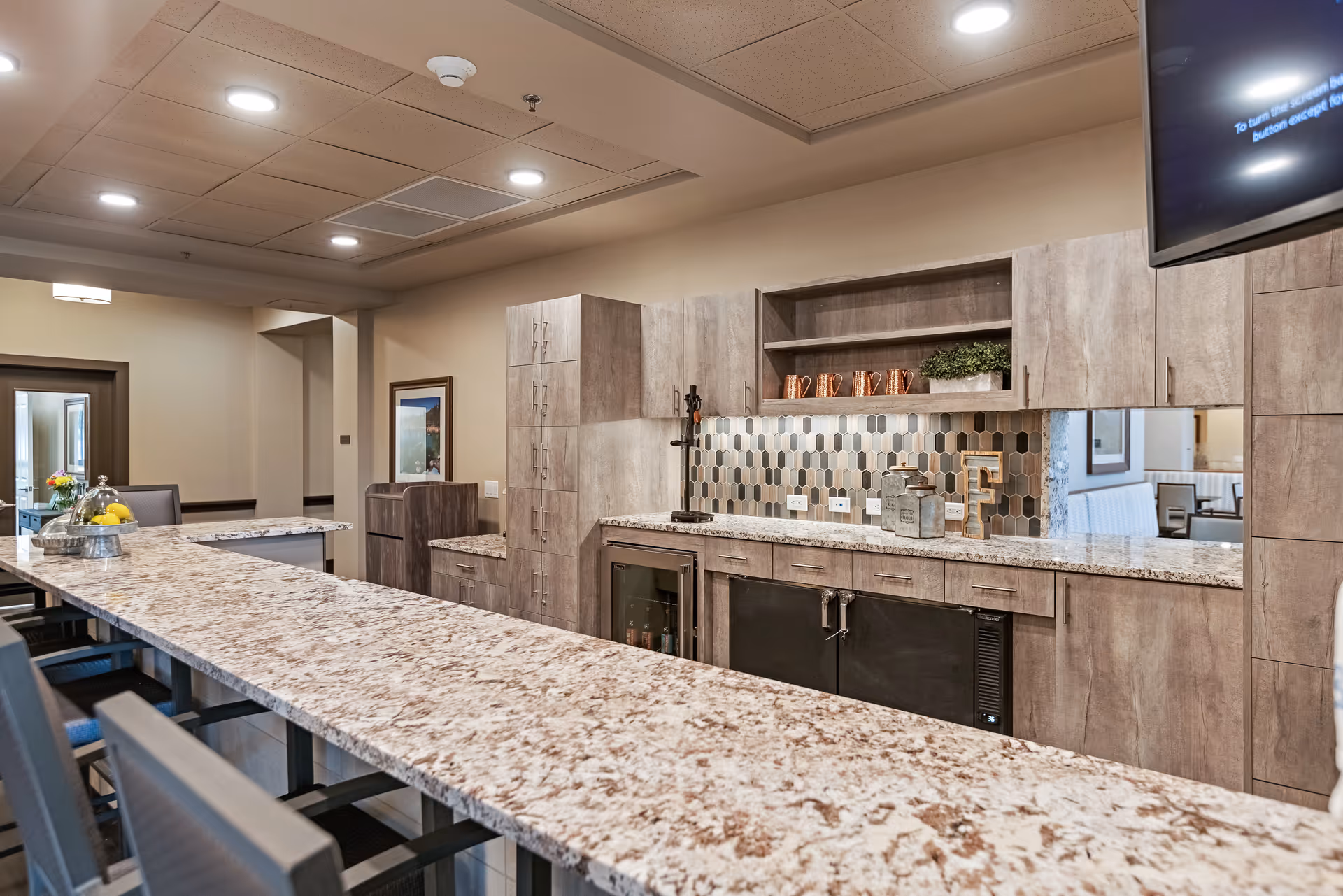 Interior view of a modern kitchen area in a senior living facility with a long granite countertop, wooden cabinets, a tiled backsplash, and bar stools lined up along the counter. The space is well-lit with recessed ceiling lights and has decorative items on the countertop and shelves.
