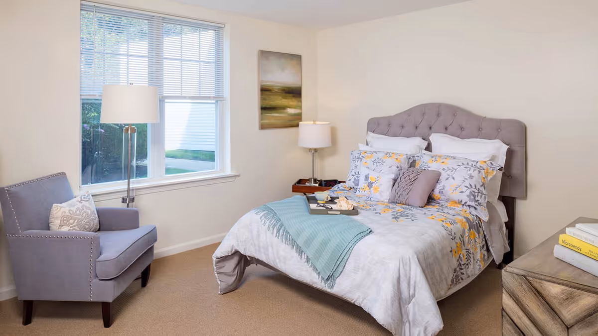 A cozy bedroom with a bed featuring a tufted gray headboard, floral bedding with yellow and gray accents, and a teal throw blanket. There is a gray armchair with a decorative pillow near a window with blinds. Two lamps, one floor and one table lamp, provide lighting. A wooden nightstand and dresser with books are also visible, along with a landscape painting on the wall.