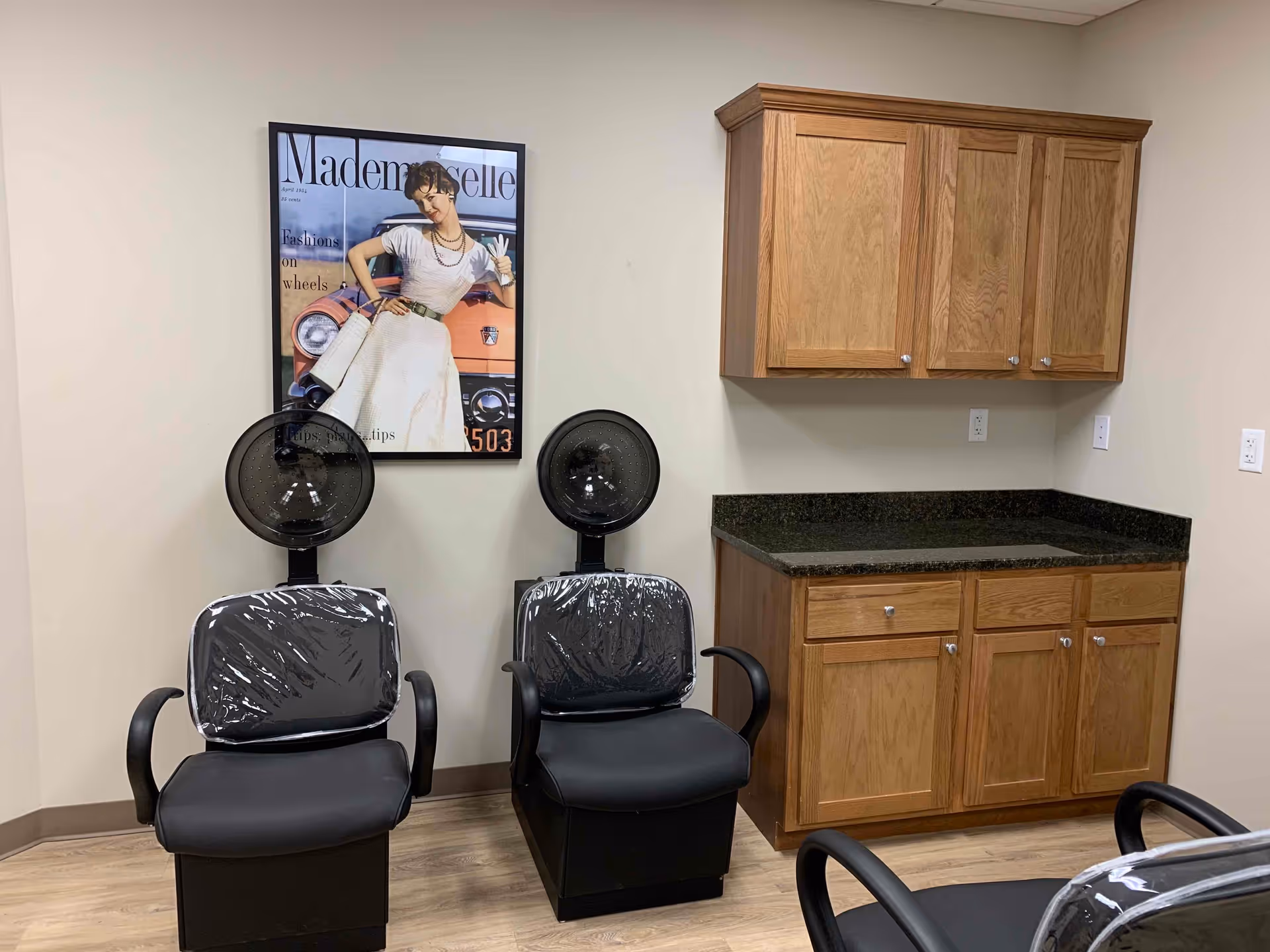 Interior room with two black salon hair dryer chairs covered in plastic, wooden cabinets with a black countertop, and a framed vintage-style poster of a woman in a white dress standing in front of a car on the wall.
