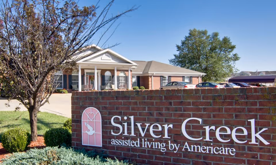Exterior view of Silver Creek assisted living facility showing a brick sign with the facility name and a building with a porch and columns in the background under a clear blue sky.