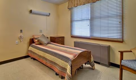 Small single-occupancy bedroom with a single bed covered by a striped bedspread, a bedside table, window with blinds, and a chair.