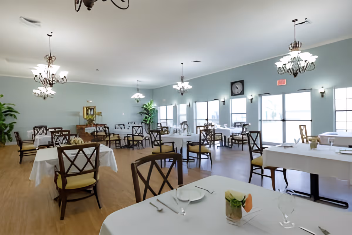 Spacious dining room with multiple set tables and wooden chairs, chandeliers, and large windows letting in natural light.