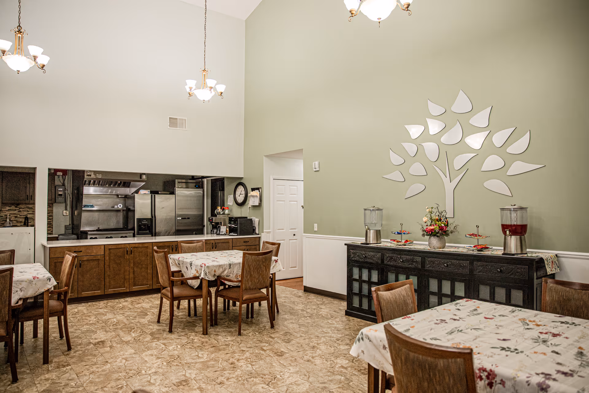 Interior view of a dining area in a senior living facility with several tables covered with floral tablecloths and wooden chairs. The room has a high ceiling with hanging light fixtures, a kitchen area with stainless steel appliances and wooden cabinets, and a decorative wall piece shaped like a tree above a black sideboard holding beverage dispensers and a flower arrangement.