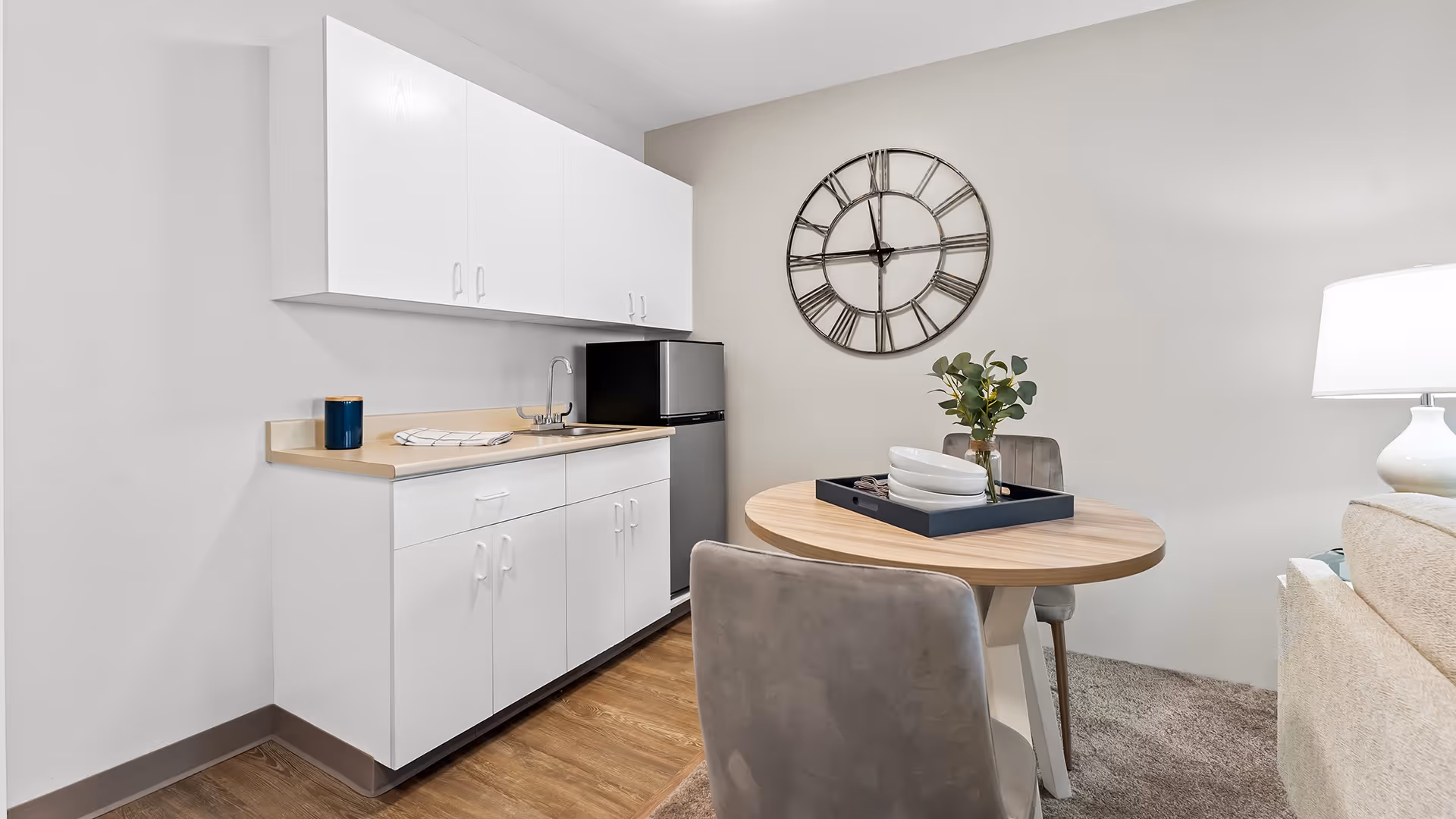 A small kitchenette area with white cabinets, a countertop with a sink, and a compact refrigerator. Next to it is a round wooden table with two gray upholstered chairs. On the table is a decorative tray with plates and a vase with green foliage. A large wall clock with Roman numerals hangs on the wall above the table. A white lamp and part of a beige sofa are visible on the right side.
