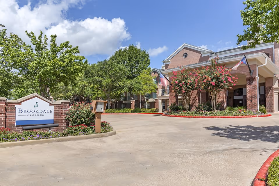 Exterior view of Brookdale First Colony senior living facility showing the entrance driveway, a brick sign with the facility name, landscaped greenery, and a building with brick columns and flags under a partly cloudy blue sky.