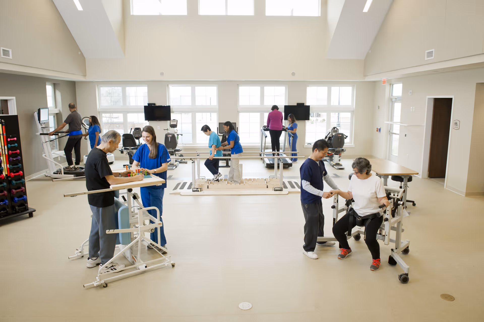 A spacious rehabilitation or physical therapy room with several elderly individuals engaged in various exercises and activities assisted by healthcare professionals. The room has large windows letting in natural light, exercise equipment including treadmills, parallel bars, and weights, and a bright, clean environment.