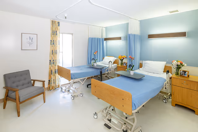 A bright healthcare facility room with two hospital beds covered in blue sheets, each with a white pillow. There are bedside tables with flowers and framed photos next to each bed. A gray cushioned chair is positioned near a window with patterned curtains. The room has light-colored walls and a clean, tiled floor.