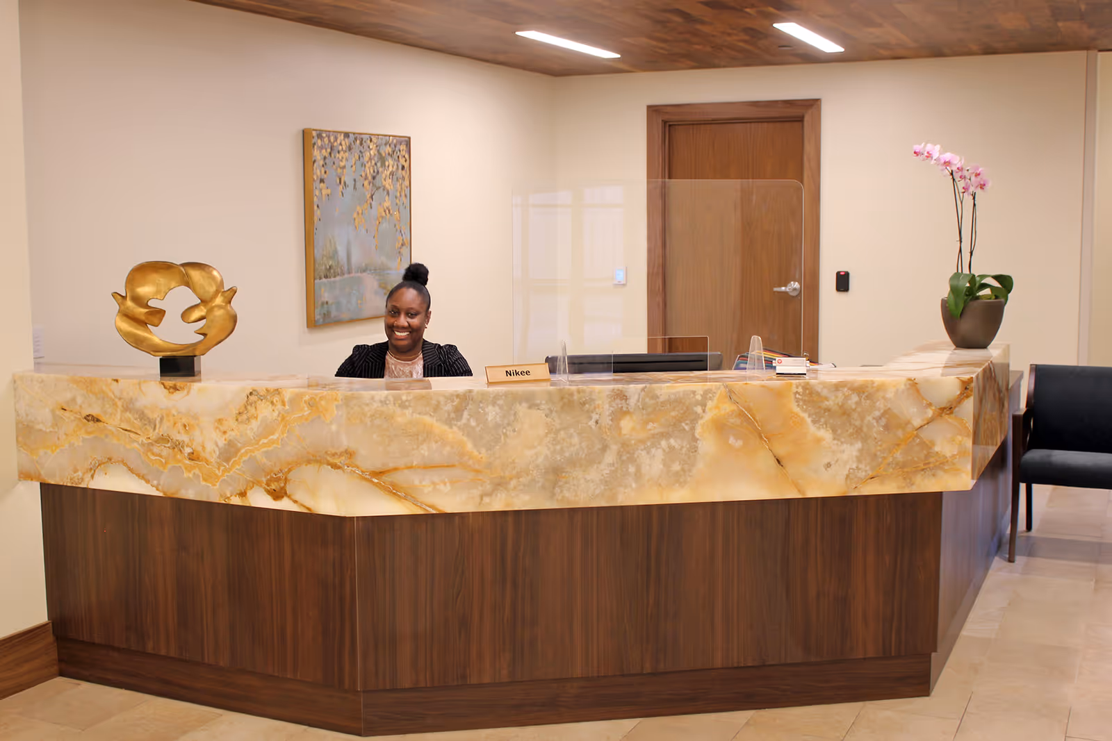 Reception area with a smiling woman behind a marble and wood front desk. The desk has a nameplate with the name Nikee, a golden decorative sculpture, and a potted orchid. A painting hangs on the wall behind the desk, and there is a wooden door and a black chair in the background.