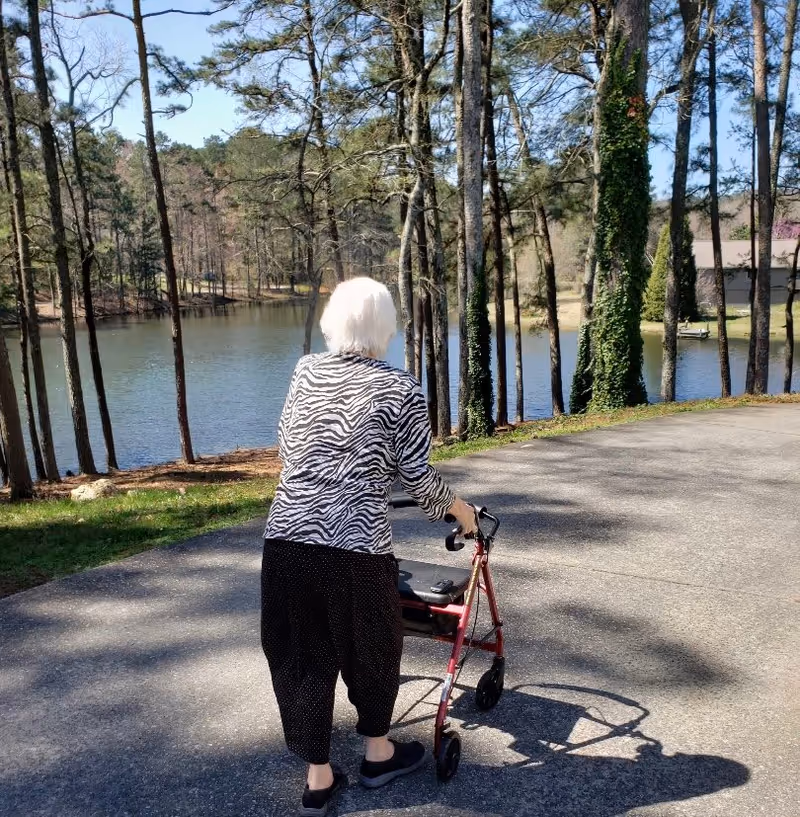 An elderly woman with white hair using a red walker is walking on a paved path near a lake surrounded by trees. The woman is wearing a zebra-striped top and black pants, and the scene is outdoors on a sunny day.