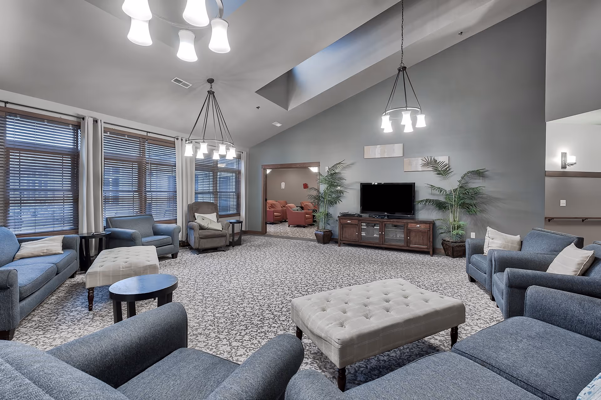 Spacious living room area with multiple blue upholstered armchairs and sofas arranged around two tufted ottomans. The room features large windows with wooden blinds and white curtains, a patterned carpet, two hanging light fixtures, a flat-screen TV on a wooden media console, and two potted plants flanking the TV. The walls are painted gray, and there is an open doorway leading to another room with red chairs.
