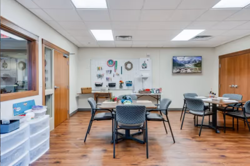 A well-lit activity room in a senior living facility with wooden floors, several tables and chairs arranged for group activities, and a pegboard on the wall holding various craft supplies. There is a framed landscape picture on the wall and wooden doors on either side of the room.
