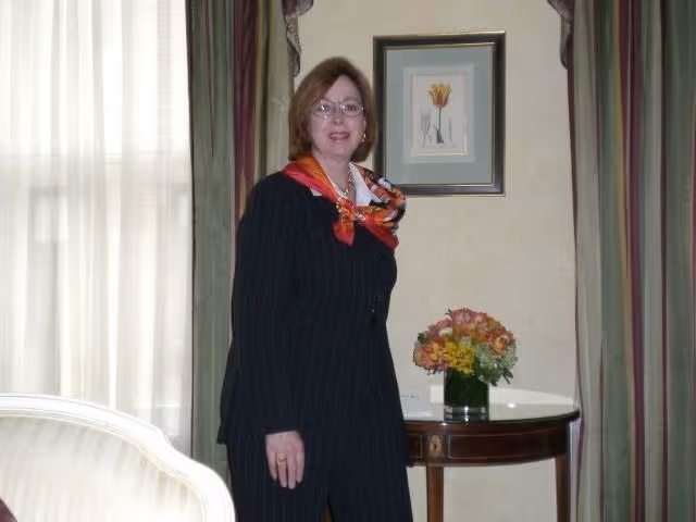 A woman wearing glasses, a dark pinstripe suit, and a colorful scarf stands in a room with a framed flower picture on the wall, a round wooden table with a vase of flowers, and curtains on the window.