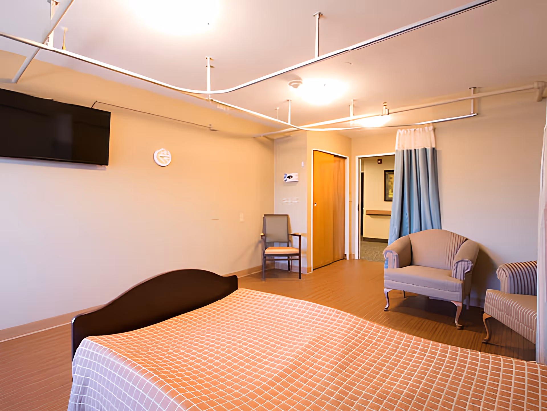 A simple bedroom in a senior living facility with a bed covered in a checkered orange and white blanket, two armchairs, a chair near the wall, a wall-mounted TV, and a ceiling-mounted curtain track system.