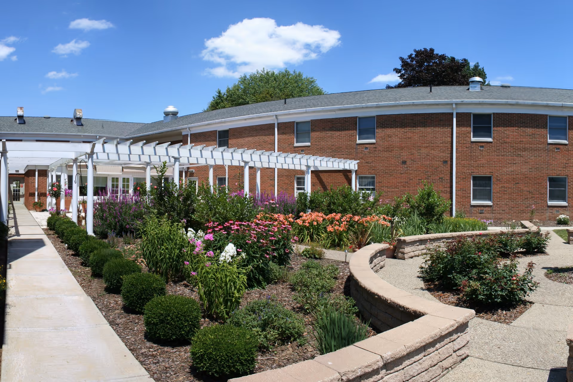 Outdoor garden area at a senior living facility with a variety of colorful flowers and shrubs, a curved stone retaining wall, a paved walkway, and a white pergola attached to a red brick building under a blue sky with a few clouds.