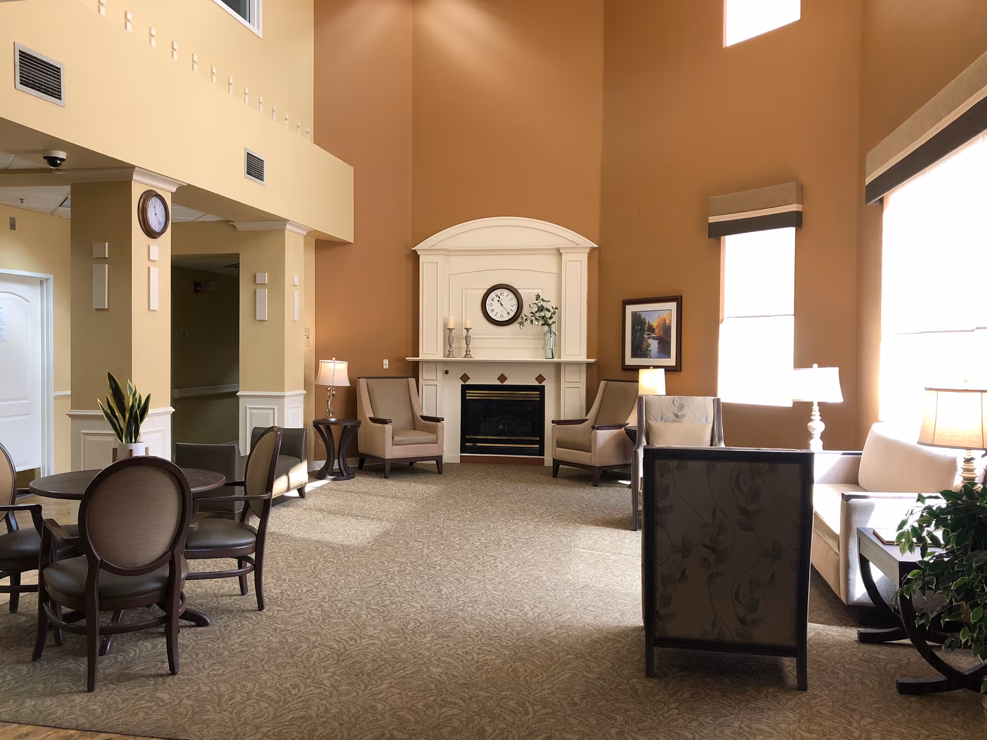A spacious and well-lit living room area in Marycrest Assisted Living featuring a fireplace with a clock and decorative items on the mantel. The room has several armchairs, a sofa, side tables with lamps, a round table with chairs, large windows with blinds, and a potted plant. The walls are painted in warm tones and the carpet has a subtle pattern.