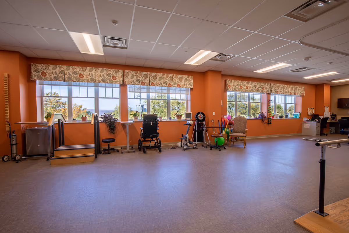 A spacious rehabilitation room with large windows covered by floral valances, allowing natural light to fill the space. The room has various physical therapy equipment including a wheelchair, exercise bike, parallel bars, and therapy balls. There are also chairs and a desk area in the background. The walls are painted orange and the floor is covered with a smooth, light-colored material.