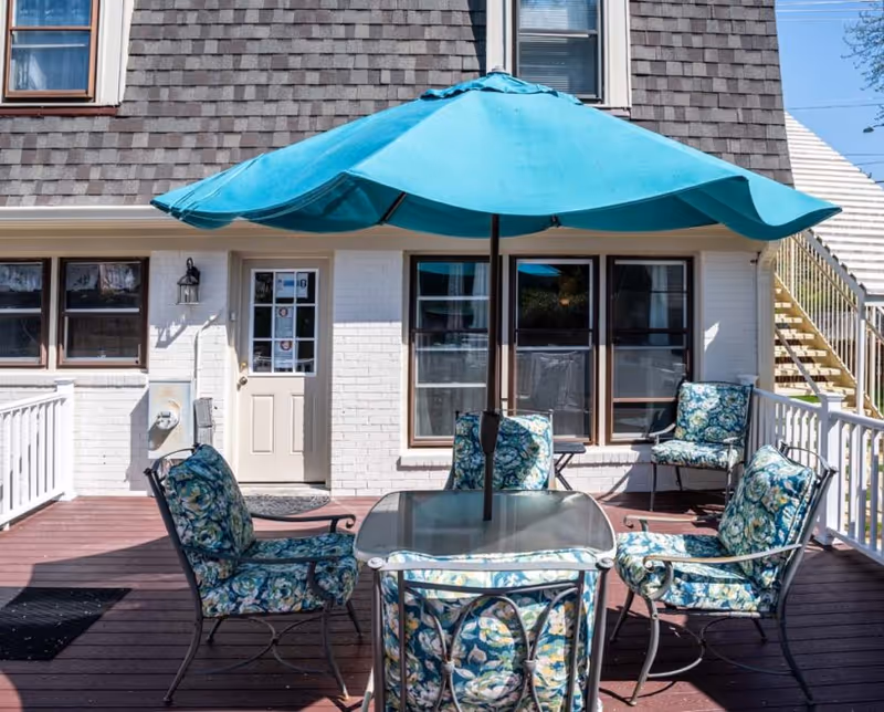 Outdoor patio area with a glass-top table surrounded by four cushioned chairs featuring floral patterns. A large blue umbrella is positioned in the center of the table. The patio is attached to a building with white brick walls and brown shingle siding on the upper floor. There are windows and a door leading inside, and a staircase is visible on the right side of the building.