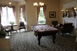 A carpeted common room with a pool table centered, upholstered chairs, a fireplace, and large bay windows letting in natural light.