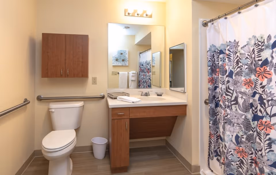 A clean bathroom with a white toilet, wooden cabinet above it, and a wooden vanity with a sink and mirror. There are grab bars on the walls near the toilet and shower. The shower has a curtain with a colorful floral pattern. A small trash bin is placed on the floor next to the toilet.