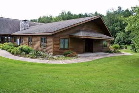 A single-story wooden building with a curved walkway, landscaped lawn, and surrounding trees.