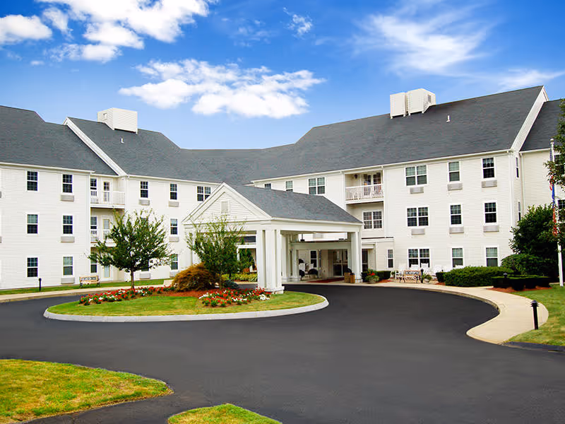 Front exterior of a white multi-story senior living building with a covered porte-cochère, circular driveway, and landscaped island.