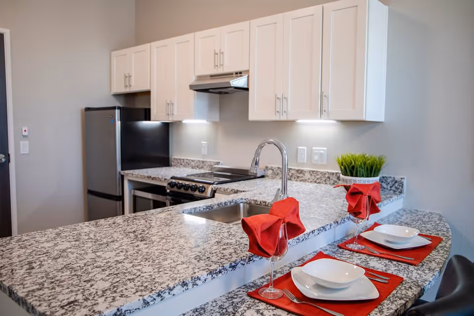 Modern kitchen with white cabinets, granite countertops, a stainless steel refrigerator, stove, and sink. Two place settings with white plates, red napkins in wine glasses, and silverware are arranged on the countertop. A small green plant is placed near the corner of the counter.
