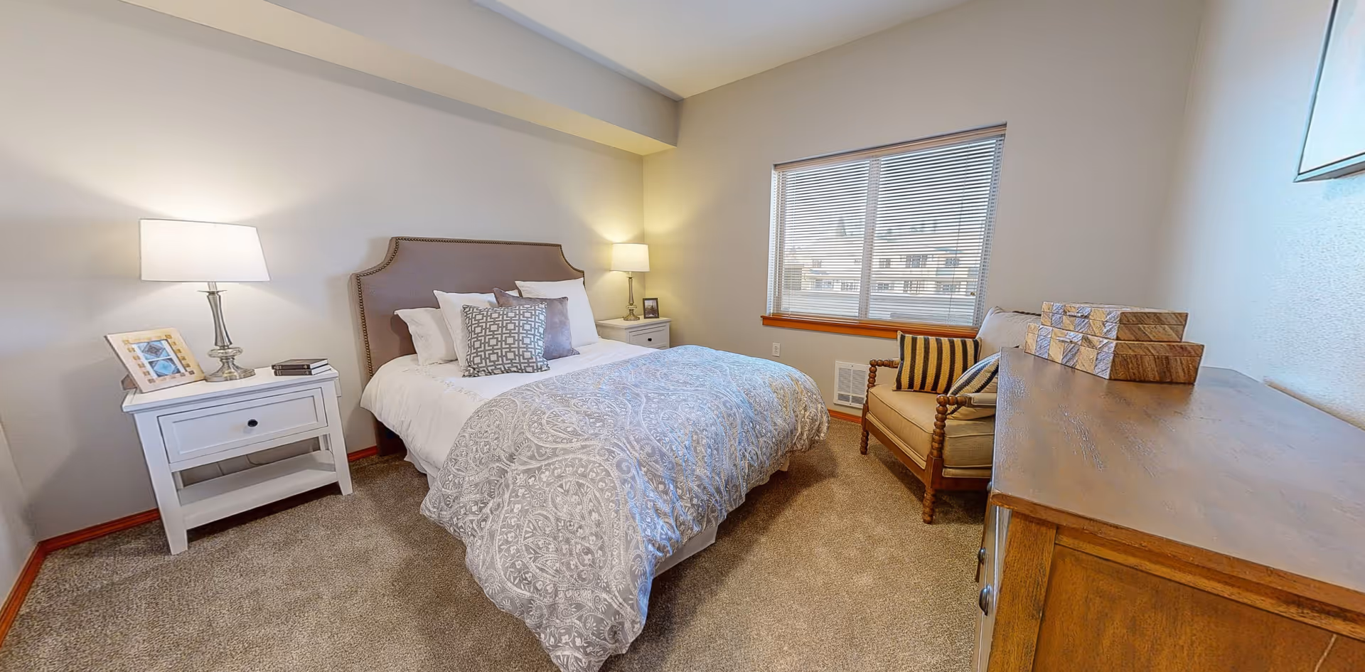 A cozy bedroom with a bed featuring a patterned gray and white comforter and multiple pillows. There are two white nightstands on either side of the bed, each with a lamp and decorative items. A window with blinds lets in natural light, and a cushioned armchair with striped pillows is placed near the window. A wooden dresser with decorative boxes is visible in the foreground.