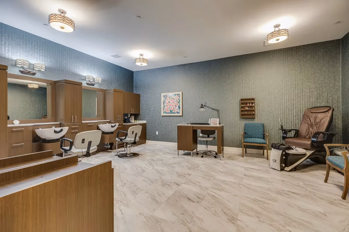 A spacious salon room with three styling stations featuring mirrors, sinks, and chairs on the left side. On the right side, there is a manicure table with a chair, a blue armchair, and a brown pedicure chair with a foot spa. The room has light-colored marble flooring, textured greenish-blue walls, and modern ceiling lights.