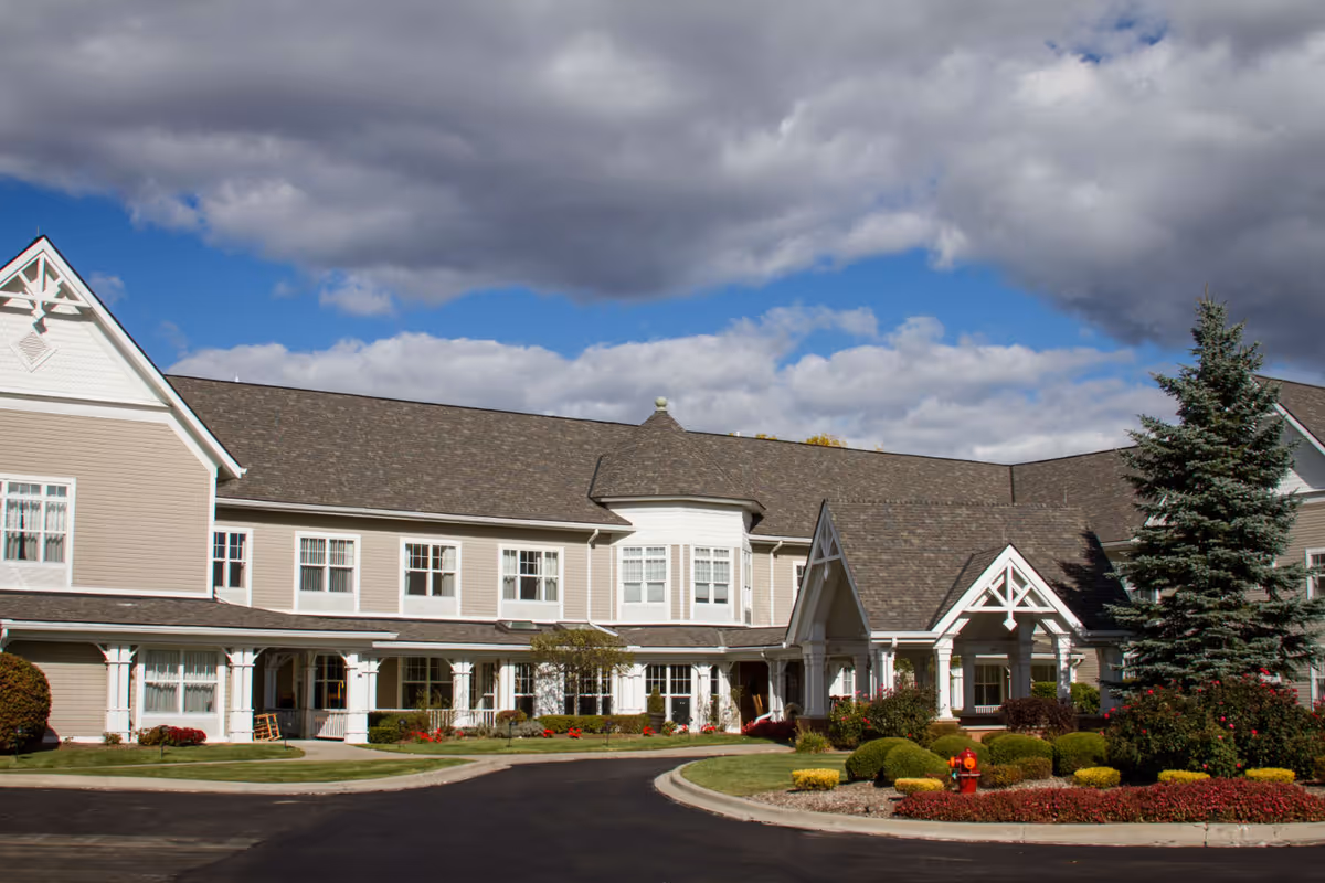 Exterior view of a large senior living facility building with beige siding, white trim, and a dark shingled roof under a partly cloudy sky. The building has multiple windows, a covered entrance, landscaped bushes, and a paved driveway in front.