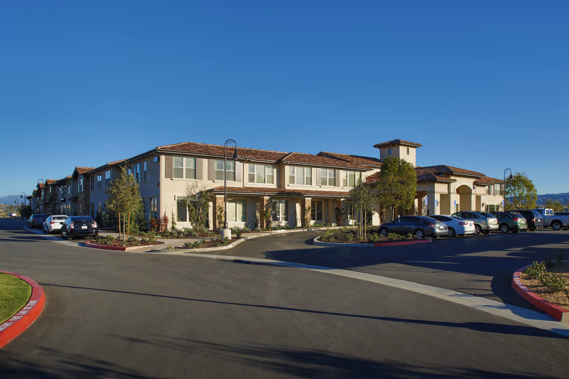 Exterior view of a two-story senior living facility building with a tiled roof, multiple windows, and a parking lot with several parked cars under a clear blue sky.