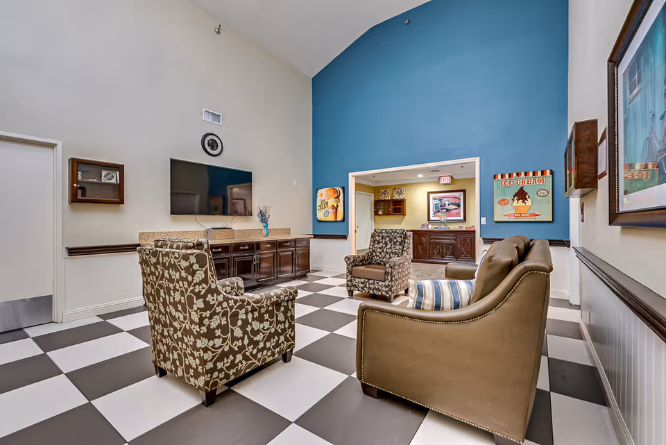 A senior living common lounge with patterned and leather armchairs around a wall-mounted TV, checkered tile floor and a blue accent wall.