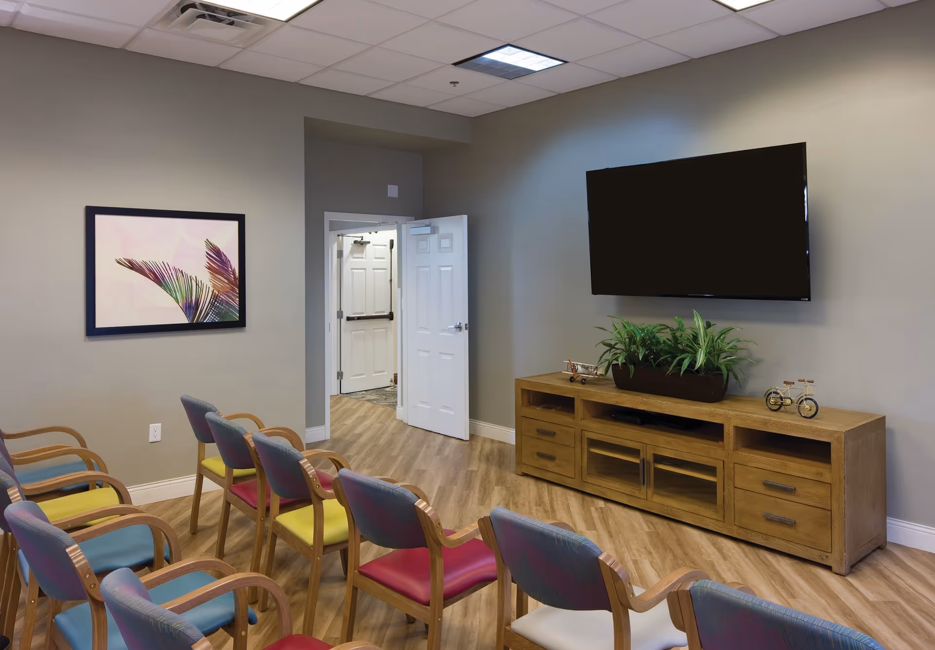 A small meeting or activity room with two rows of colorful chairs facing a large flat-screen TV mounted on a beige wall. Below the TV is a wooden cabinet with drawers and decorative items including a plant and small model bicycles. A framed artwork featuring palm leaves is hung on the left wall. The room has wood-look flooring and a white door is open, leading to another area.