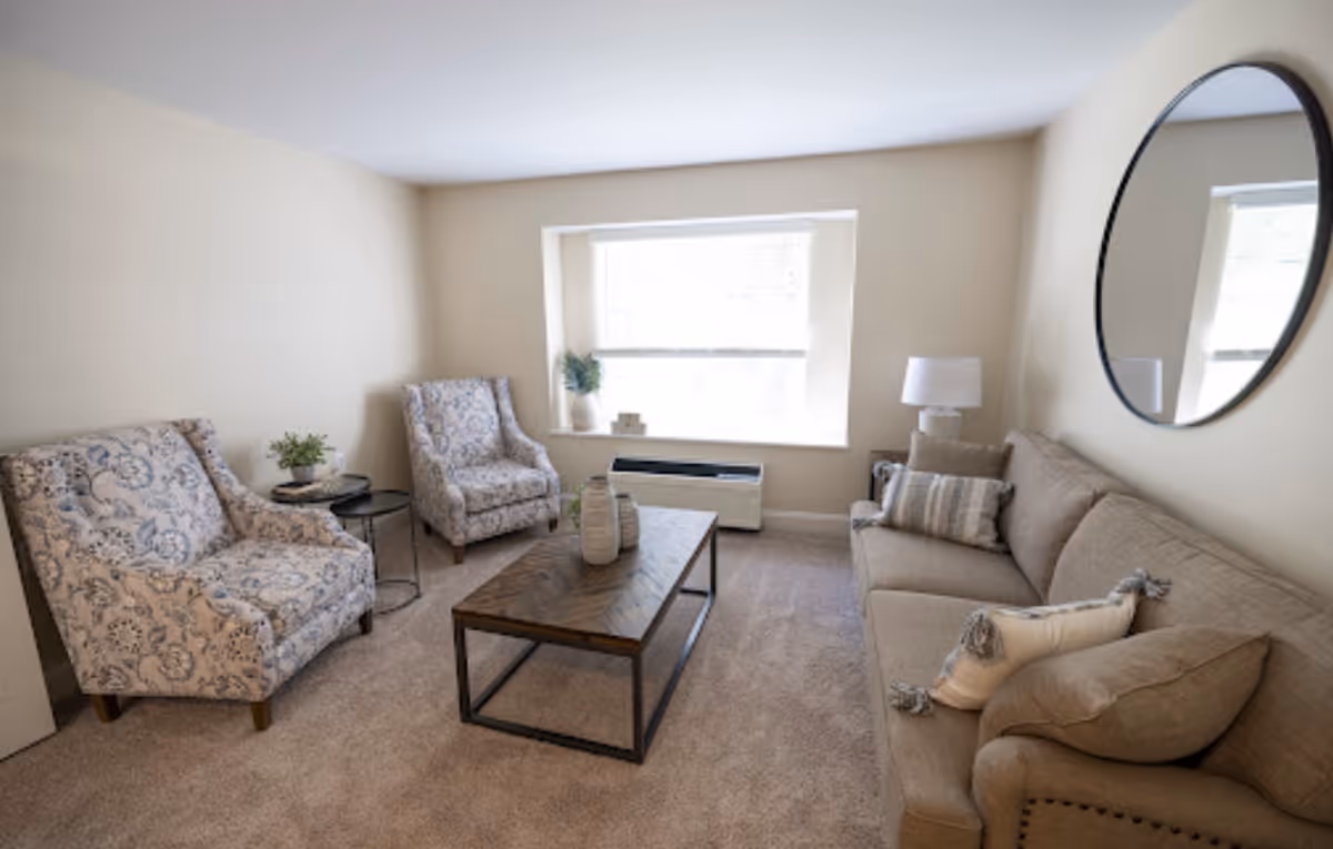 A cozy living room with a beige sofa adorned with pillows, two patterned armchairs, a wooden coffee table with decorative vases, a round mirror on the wall, a table lamp, and a window letting in natural light.