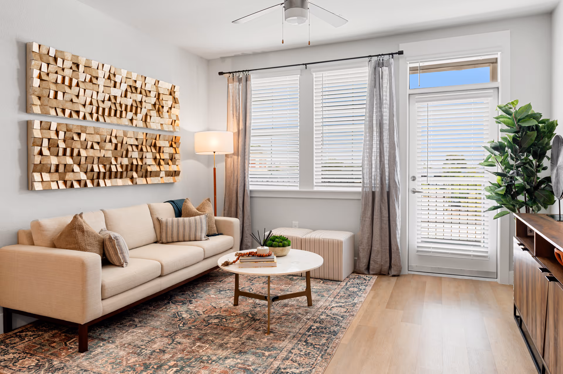 Sunlit modern living room with a beige sofa, round coffee table, textured wall art, rug, and a glass door with blinds.