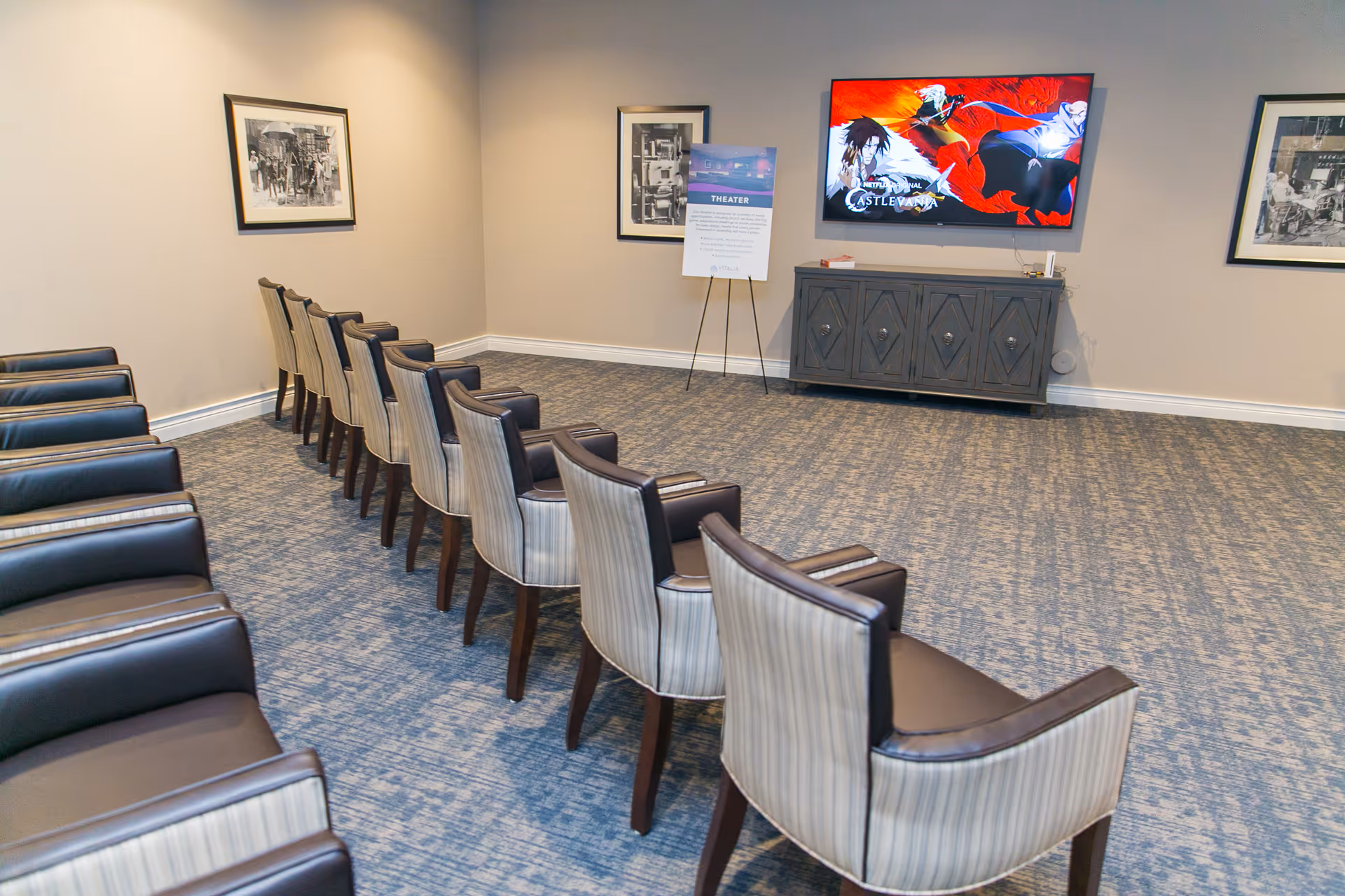 A small theater room with two rows of chairs facing a wall-mounted flat screen TV displaying an animated show. The room has carpeted flooring, beige walls, framed black and white pictures, and a cabinet below the TV. There is also a sign on a stand labeled 'THEATER' near the TV.