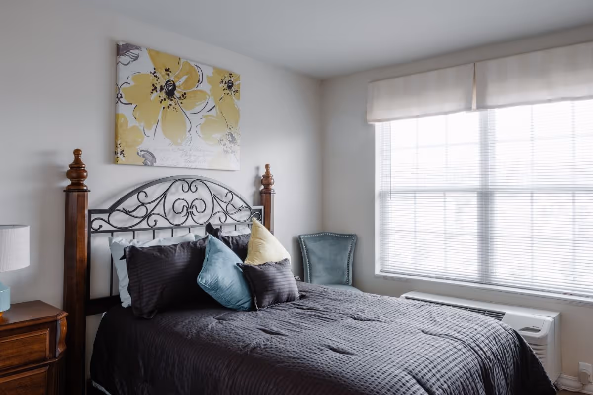 Sunlit bedroom with an ornate metal headboard, dark bedspread and colorful throw pillows beside a large window.