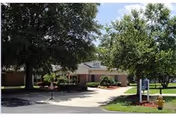 Exterior view of a single-story brick building with a driveway and landscaped greenery including trees and bushes under a partly cloudy sky.