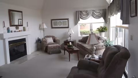A cozy living room with a fireplace on the left, a cushioned armchair in the foreground, a sofa and an armchair with a matching ottoman near the window, a coffee table with a lamp and decorative items, and large windows with draped curtains letting in natural light.