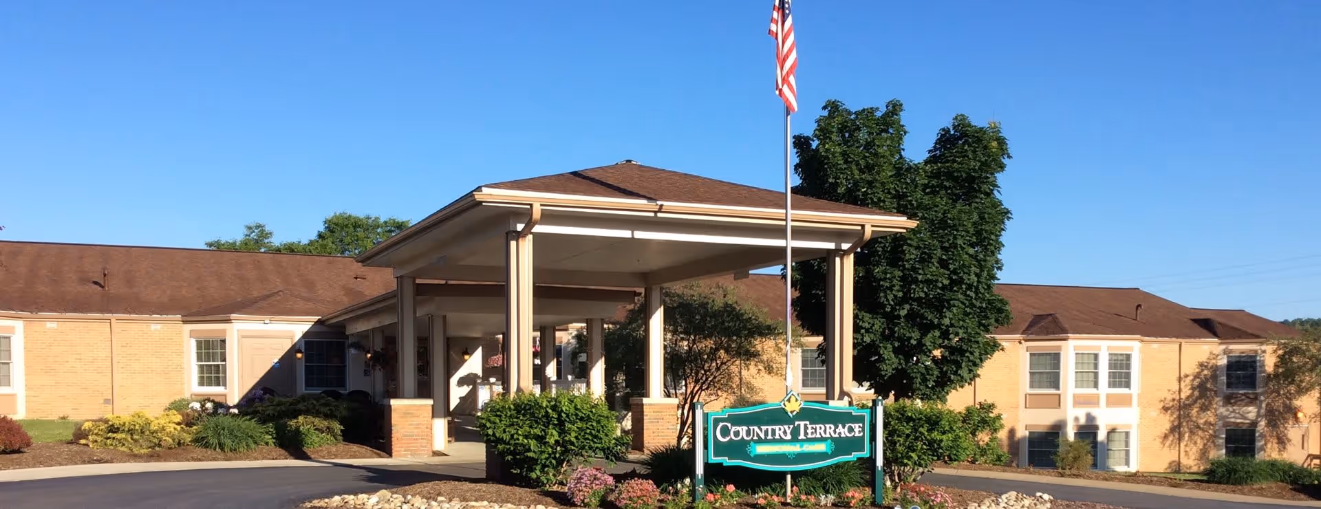 Exterior view of Country Terrace senior living facility showing a single-story building with a covered entrance, an American flag on a flagpole, landscaped bushes and flowers, and a clear blue sky.