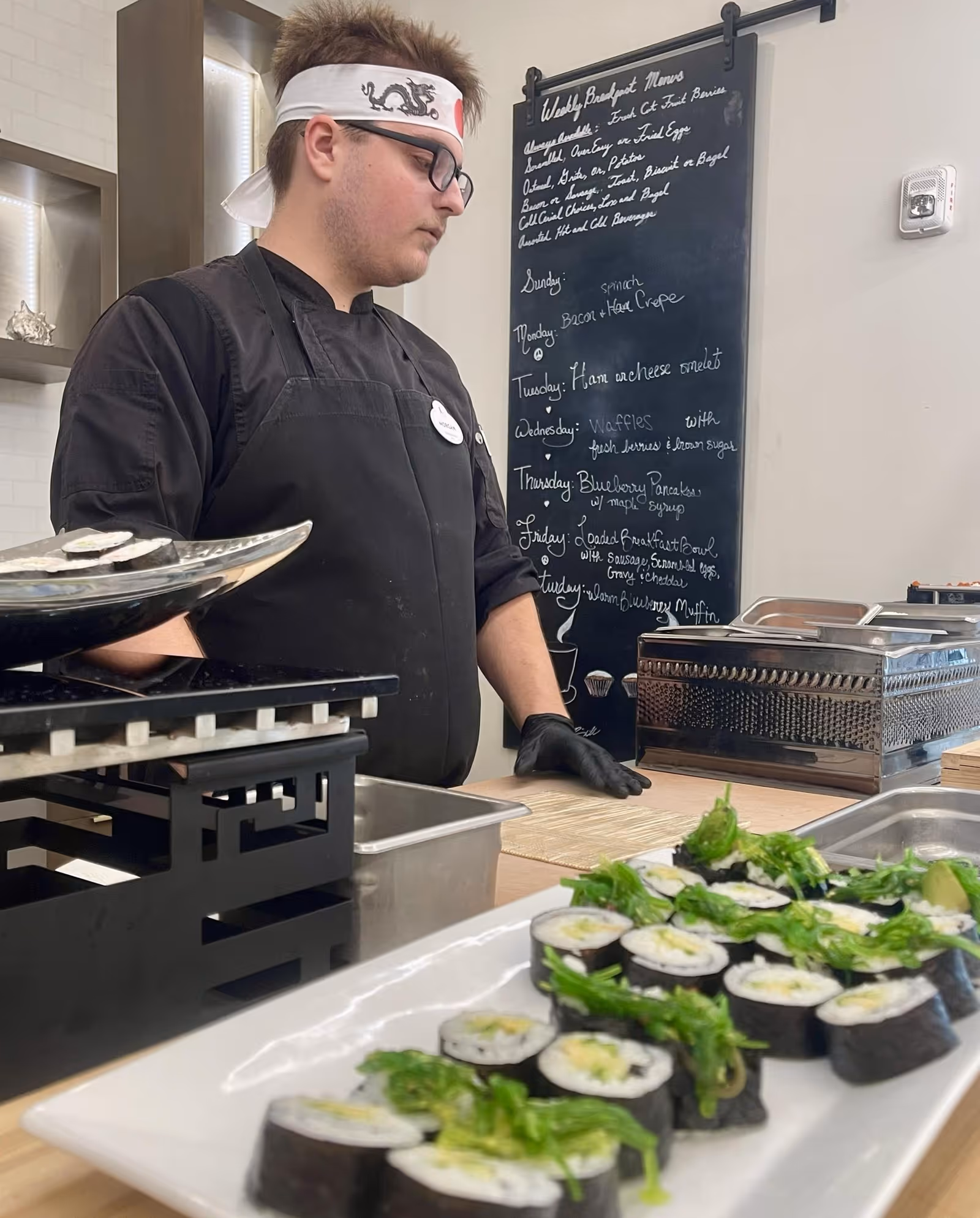A chef wearing a black apron, black gloves, glasses, and a white headband with a dragon design is preparing sushi rolls in a kitchen. In the foreground, there is a white plate with neatly arranged sushi rolls topped with green garnish. Behind the chef, a blackboard displays a weekly breakfast menu written in white chalk.