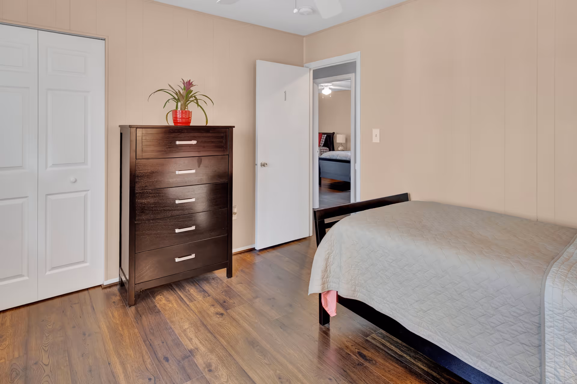 A simple bedroom with wooden flooring, beige walls, a dark wooden dresser with a red potted plant on top, a bed with a light gray quilt, and an open door showing another bedroom in the background.