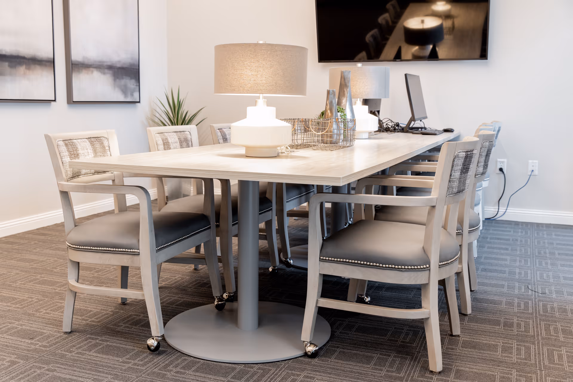 A modern conference or meeting room with a long rectangular table surrounded by eight cushioned chairs with armrests. The table has two decorative lamps and a wire basket with small plants and decor items. A large flat-screen TV is mounted on the wall in the background, and two abstract paintings hang on the adjacent wall. The room has a carpeted floor and neutral-colored walls.