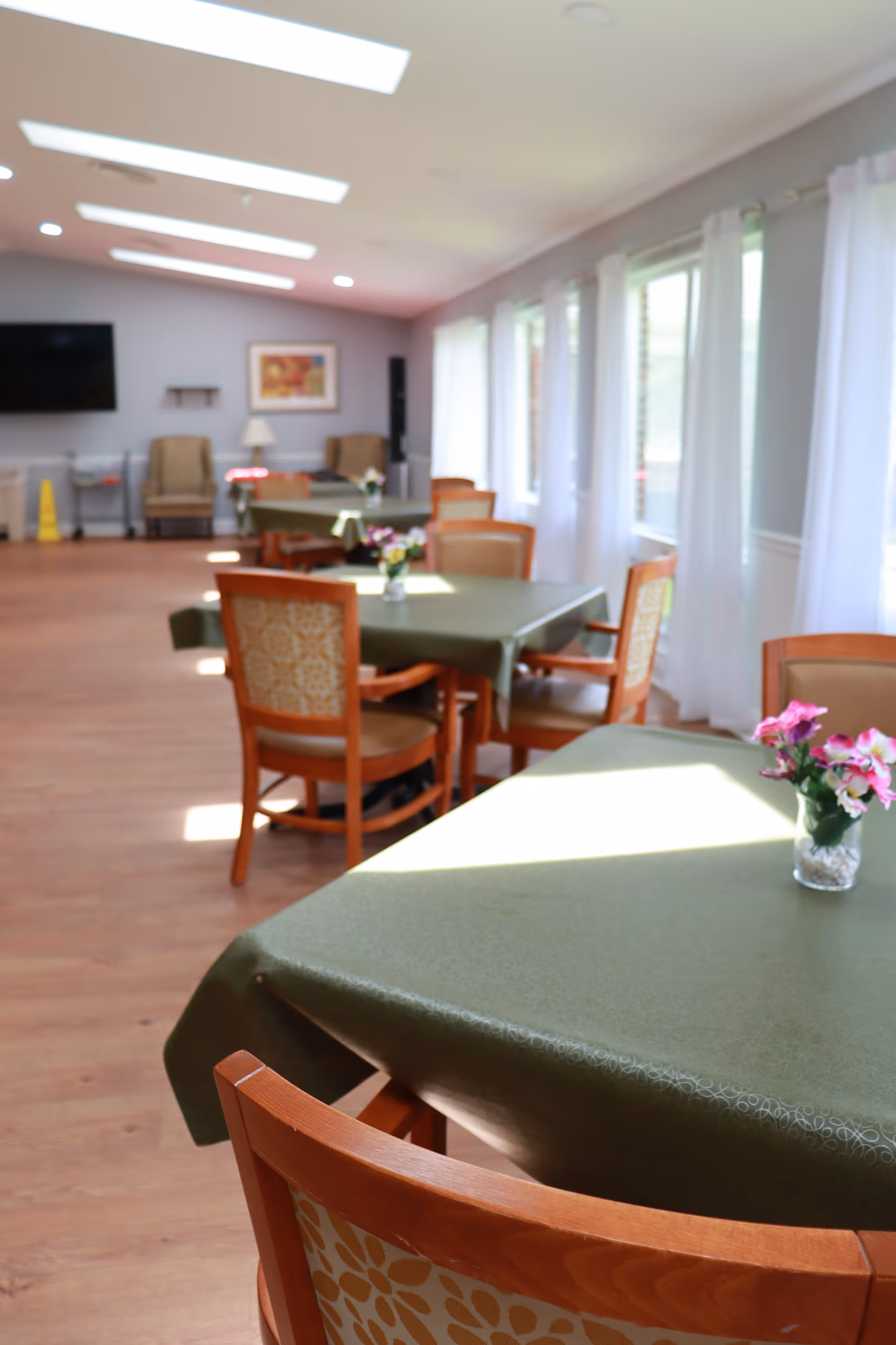 A bright dining room with several tables covered in green tablecloths, each adorned with small vases of pink flowers. Wooden chairs with patterned upholstery surround the tables. Large windows with white curtains allow natural light to fill the room. In the background, there are armchairs, a wall-mounted television, and a framed picture on the wall.