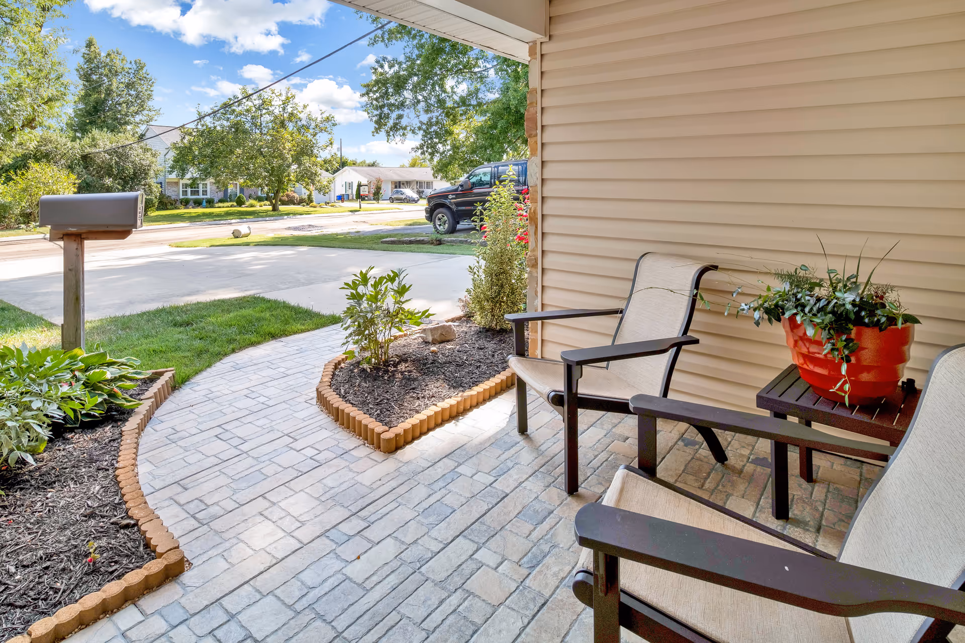 View of a covered front porch with two beige cushioned chairs and a small table holding a red flower pot with green plants. The porch has a stone tile floor and overlooks a curved paved walkway bordered by small garden beds with plants and mulch. A mailbox is visible near the street, with houses, trees, and a parked black truck in the background under a partly cloudy blue sky.