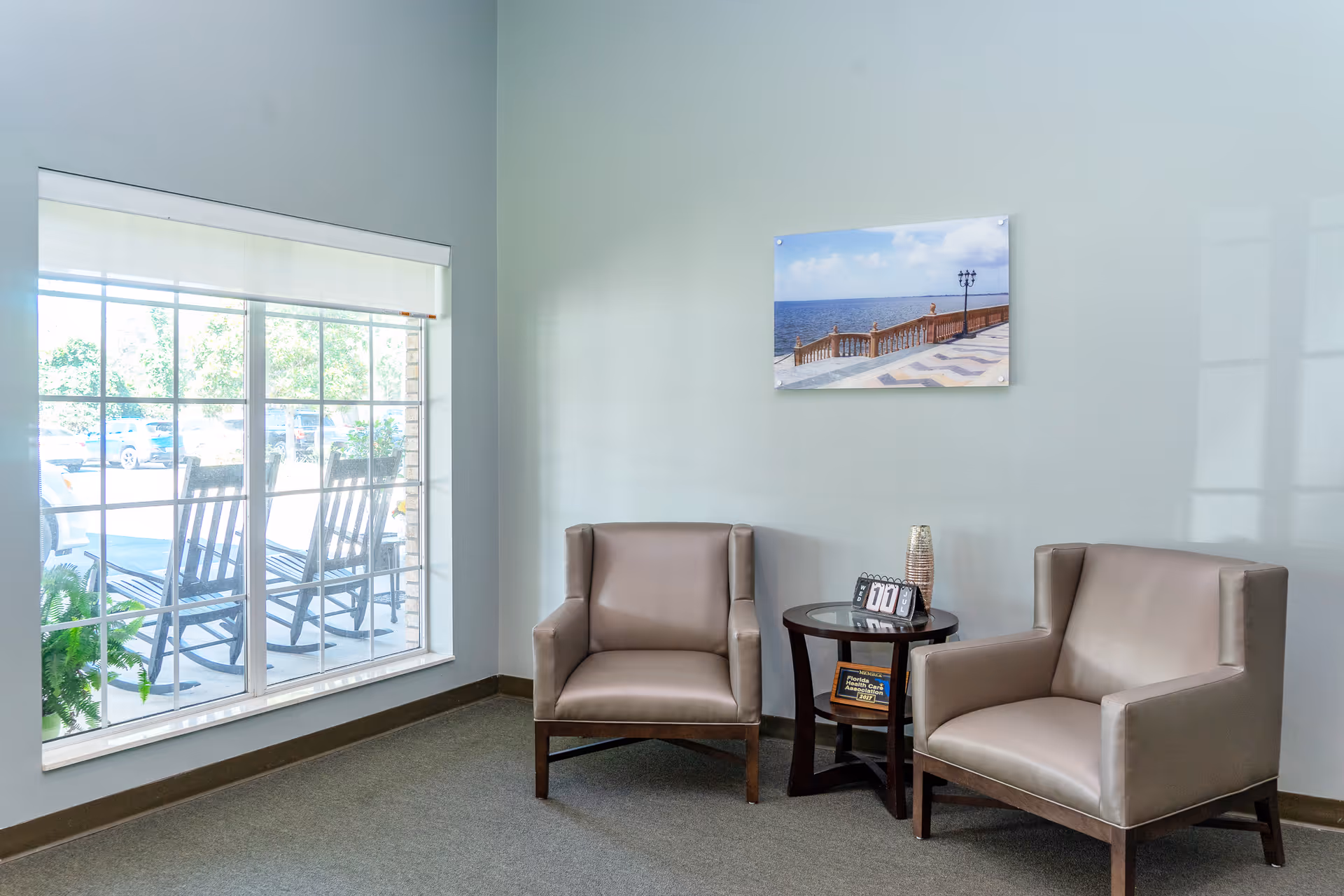 A small seating area with two beige armchairs and a round wooden side table between them. On the table, there is a decorative vase and a small calendar displaying the number 11. A large window with a view of outdoor rocking chairs and greenery is on the left side. A framed picture of a seaside promenade hangs on the light gray wall behind the chairs.