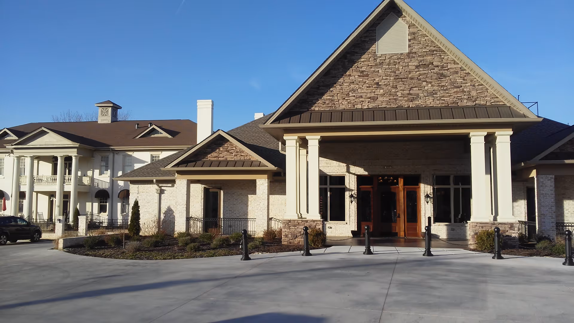 Front exterior view of a senior living facility building with a large covered entrance supported by white columns, stone and brick facade, and clear blue sky.