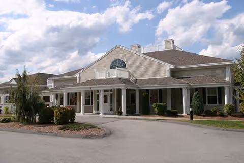 Exterior view of a single-story senior living facility building with beige siding, white columns, and a covered entrance. The building is surrounded by landscaped bushes and trees under a partly cloudy sky.